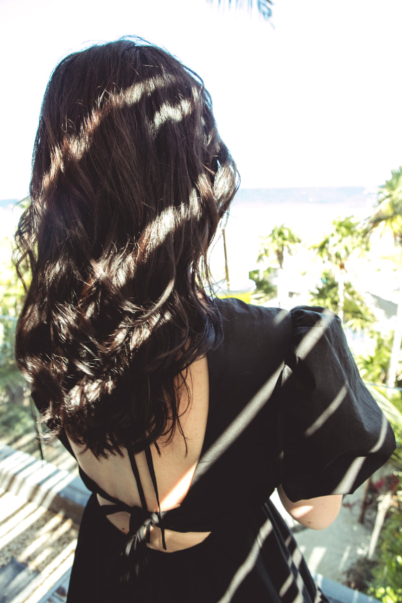 A woman viewed from behind wearing a black backless dress with short puff sleeves, standing outdoors with palm trees and a bright sky in the background. Her dark wavy hair is illuminated by dappled sunlight.